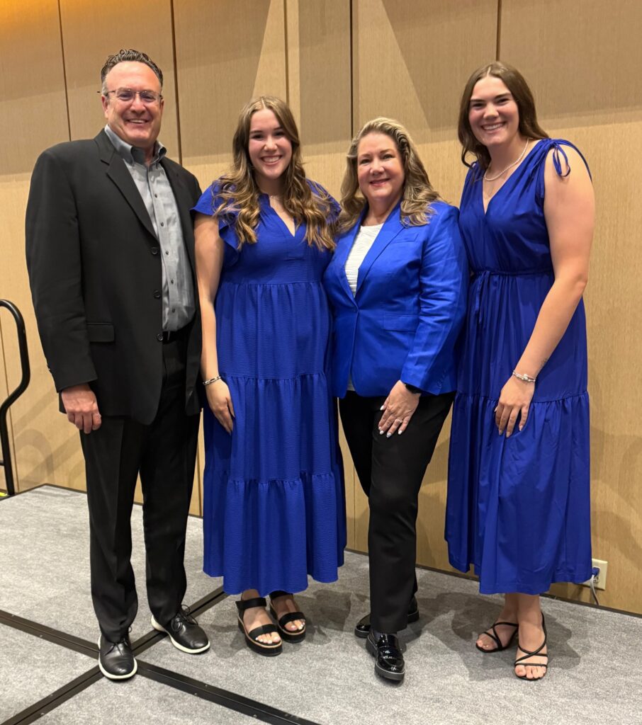 This is a posed picture of four people. Heather Collins, president of AWWA, is third from left. Her daughters are on either side of her, and her husband is far left. The women are all dressed in blue, a tribute to water, for the ceremony officially recognizing Collins as president.